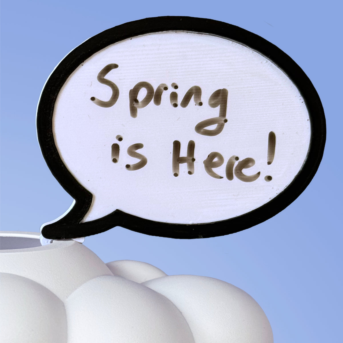 A white, cloud-shaped dry erase word bubble charm with spring is here written on  it, attached to a plant.