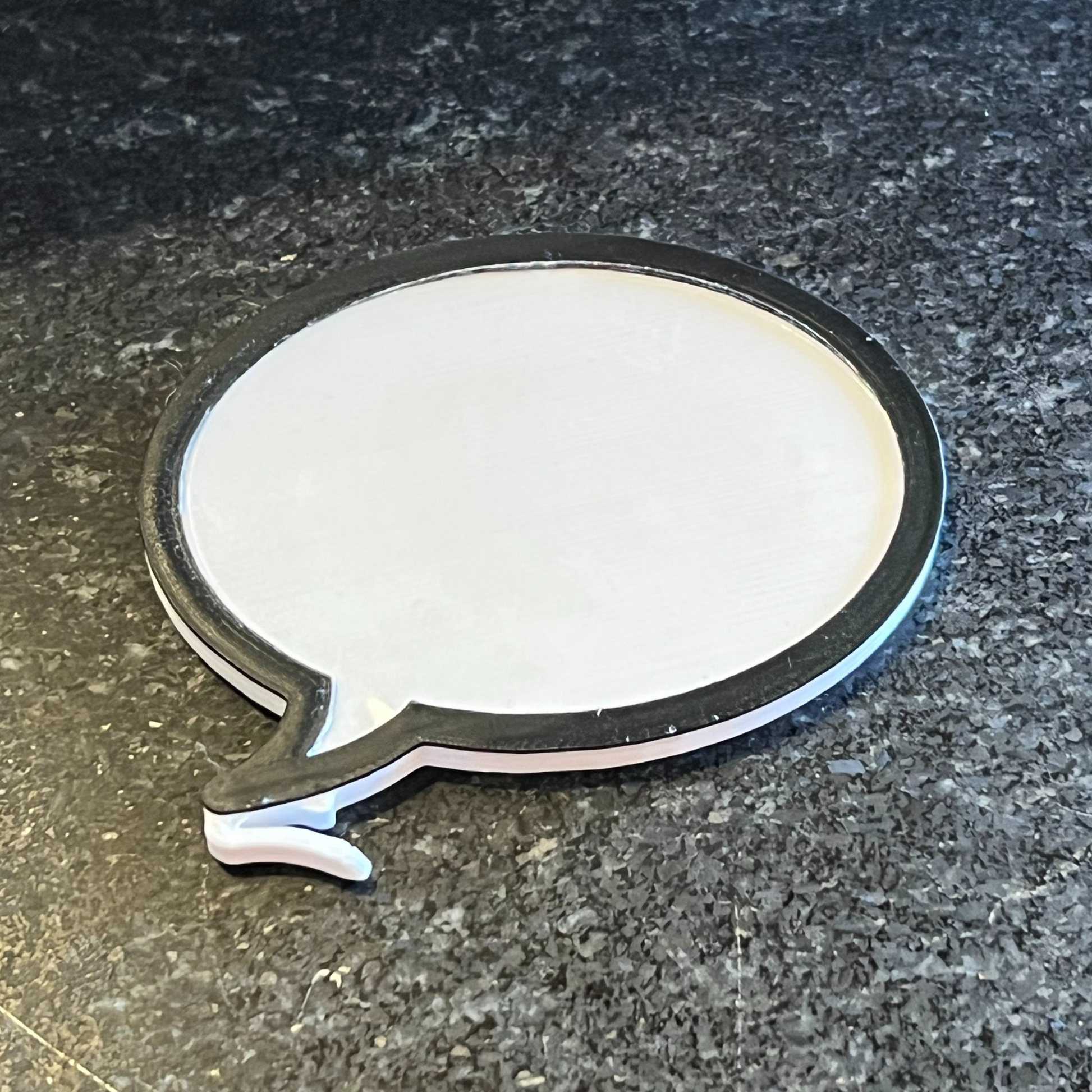This image displays a dry erase word bubble charm, designed in a Blank slate color with a speech bubble shape. The charm features a sleek, minimalist style with a smooth, white surface and a black outline that contours the bubble's form.