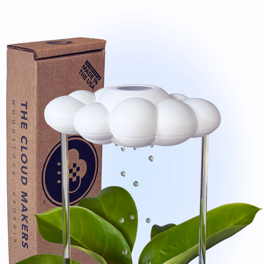 A white cloud-shaped plant watering device with water droplets, placed on two acrylic rods, against a green leafy plant backdrop with a brown box labeled 'THE CLOUD MAKERS'.