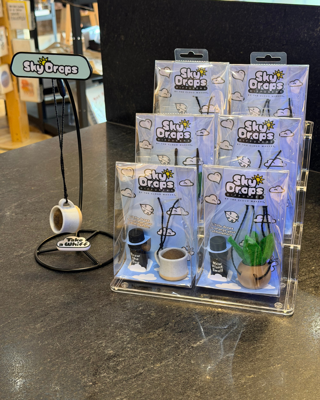 Six packaged SkyDrops coffee aair fresheners displayed in a clear acrylic stand on a dark countertop, with one air freshener hanging on a black display stand to the left.