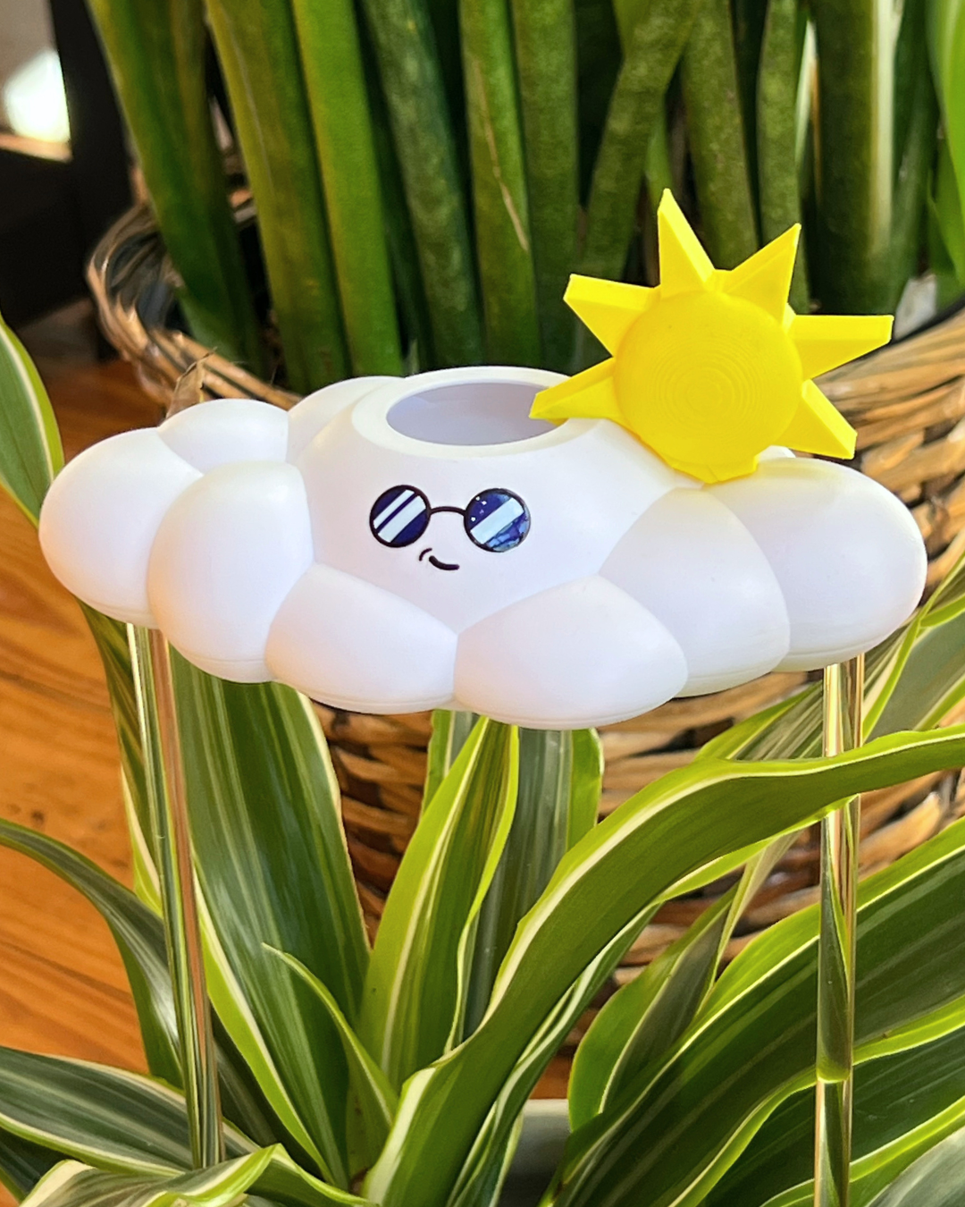 A white cloud-shaped plant waterer with a smiling face wearing blue sunglasses and a yellow sun decoration on top, surrounded by green houseplants.