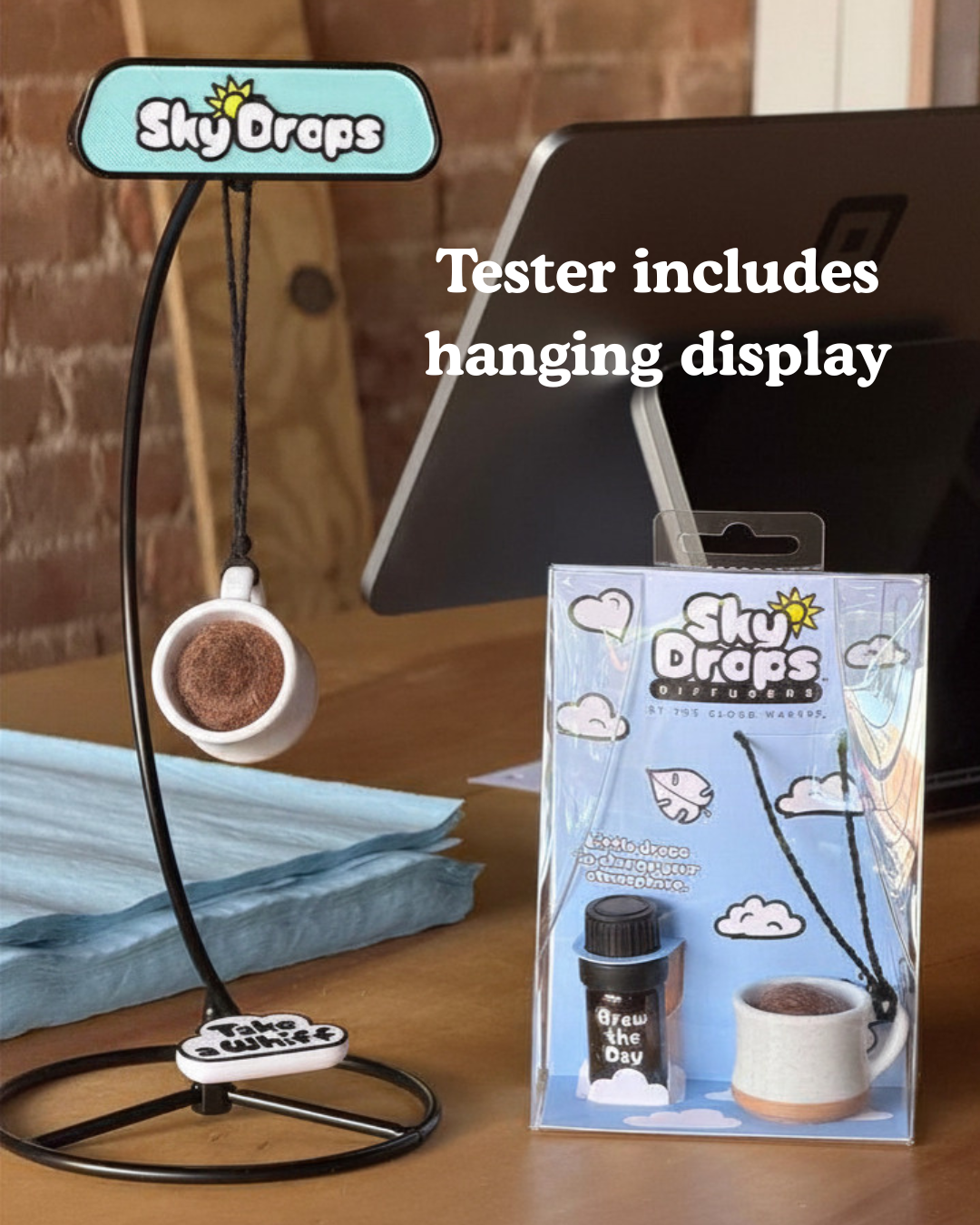 A coffee mug hanging display kit by SkyDrops called 'Brew the Day' shown with its packaging on a wooden desk in front of a laptop. The black metal stand features a light blue sign at the top and a small white mug hanging from a curved black arm.